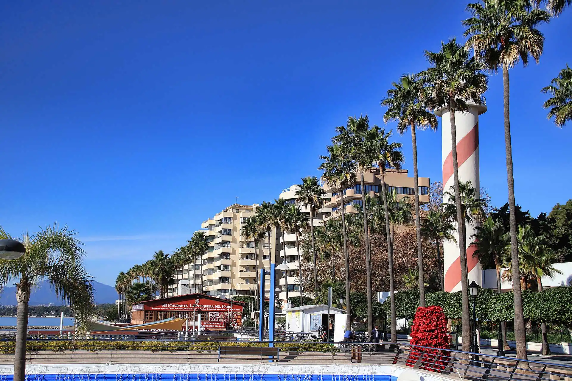View of Marbella's Paseo Marítimo with palm trees, modern buildings, and the iconic lighthouse under a clear blue sky.