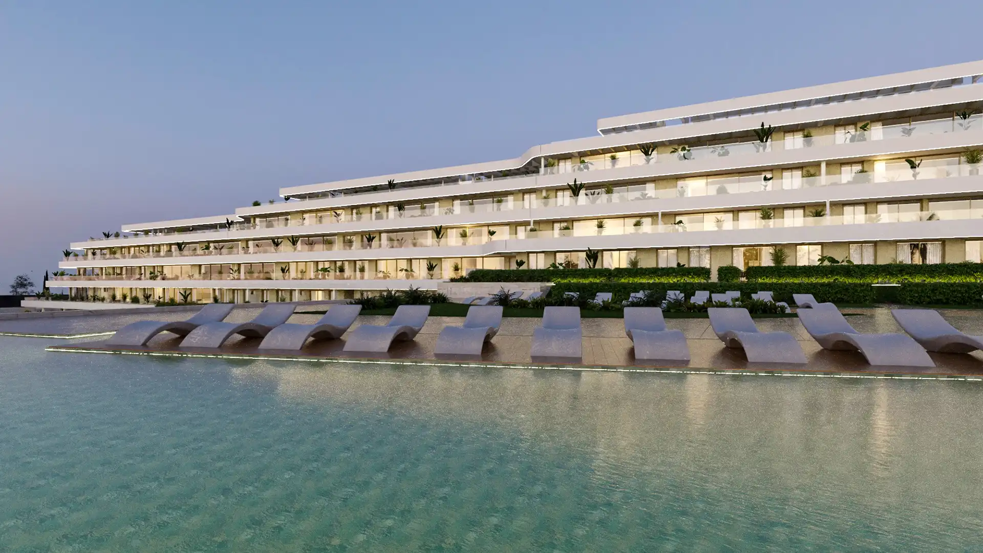 Modern white apartment building with wide terraces and sun loungers by an illuminated swimming pool at dusk in Arosa, Torrenueva, Mijas.