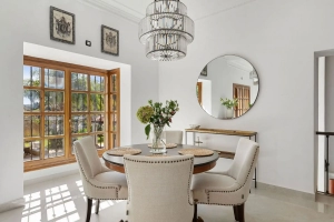 Dining area featuring a round table with upholstered chairs and a chandelier above, with a large window in the background.