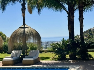 Two lounge chairs under a thatched umbrella surrounded by palm trees and greenery.