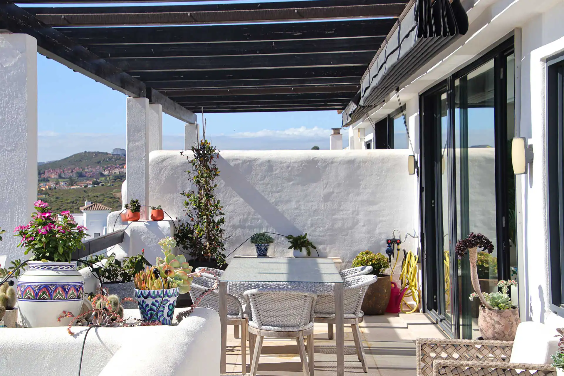 Covered outdoor terrace with dark wood pergola, wicker dining set, colorful potted plants, and panoramic mountain views in Mediterranean style property