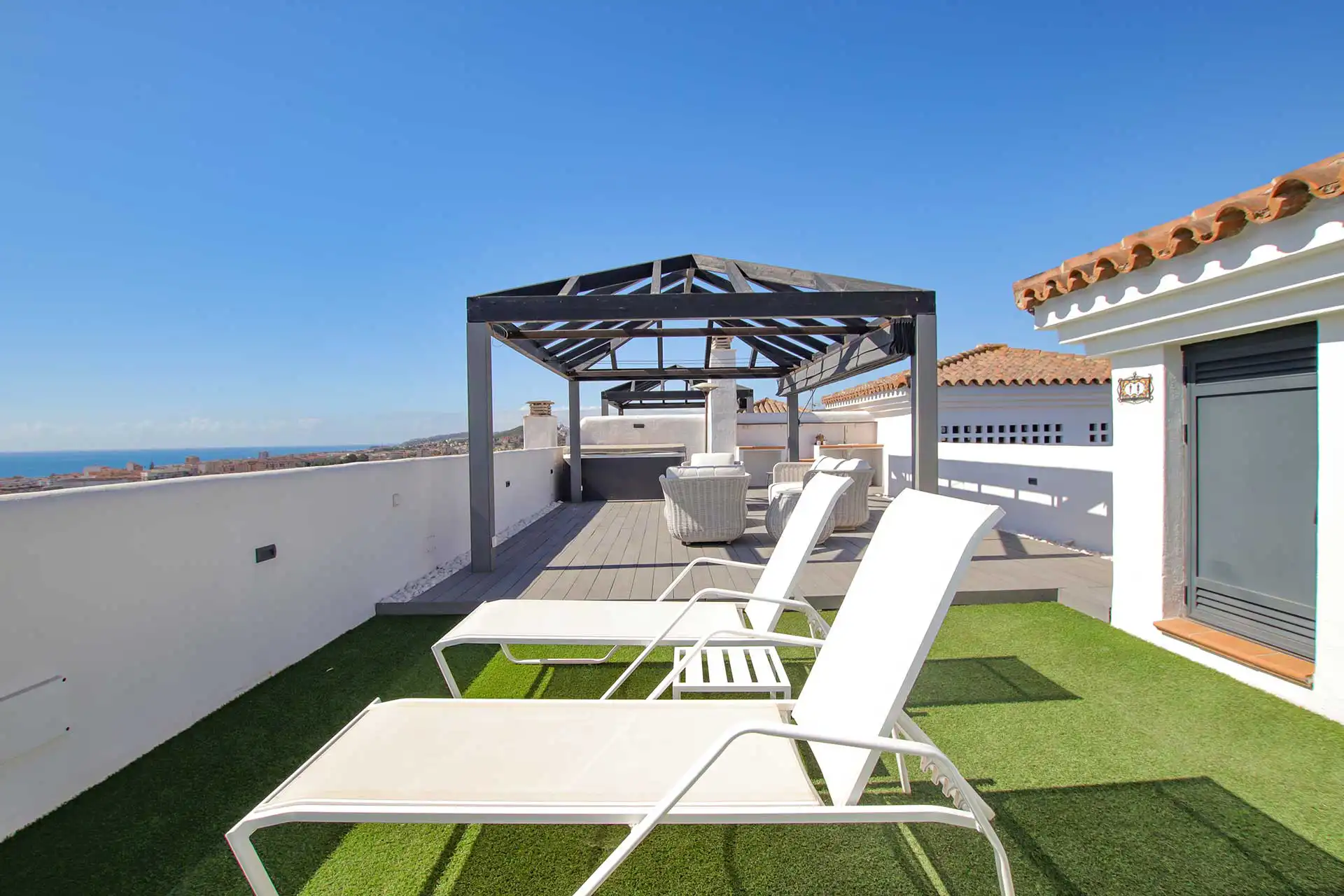 Luxury rooftop terrace with white sun loungers, artificial grass, dark metal pergola, and panoramic Mediterranean sea views