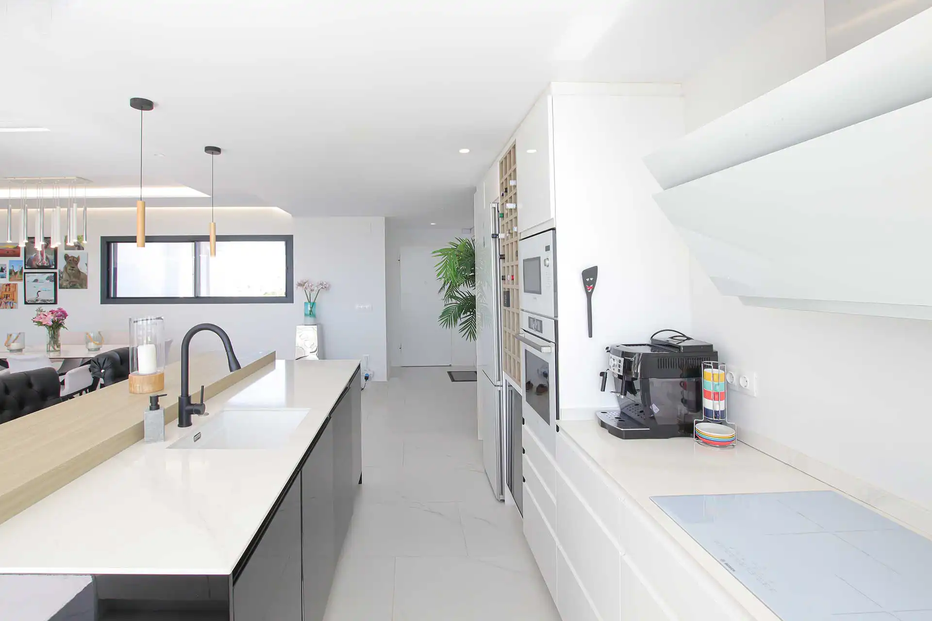 Sleek white modern kitchen featuring a large island with black faucet, built-in ovens, espresso machine, and open dining area