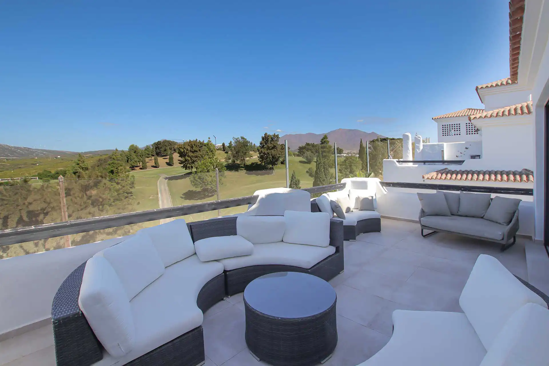 Spacious luxury rooftop terrace with curved white wicker sofa set, glass balustrade, overlooking lush golf course and mountains