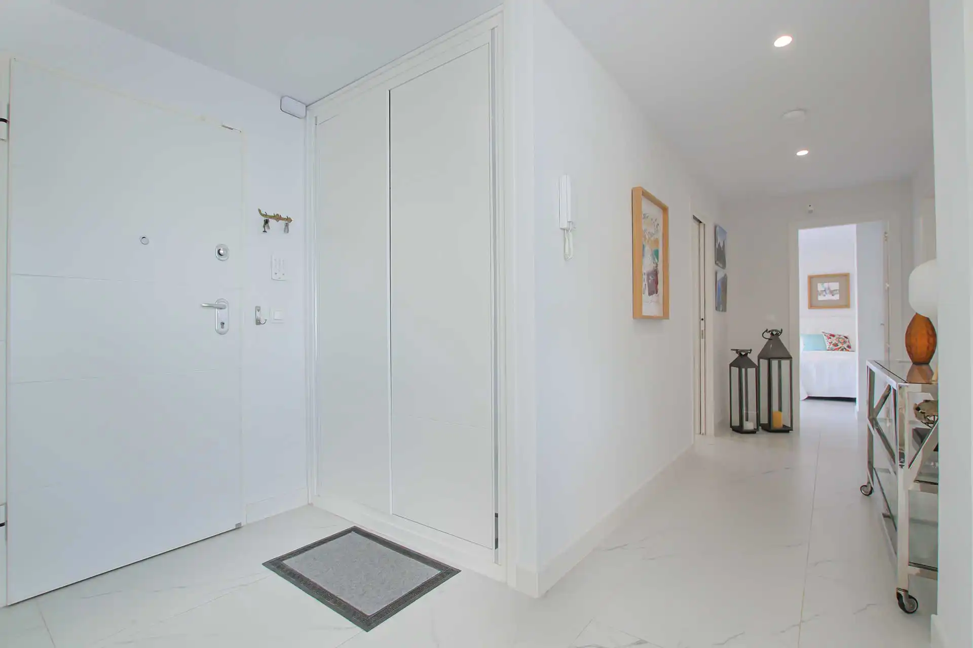 Spacious white entrance hallway with marble tile flooring, decorative lanterns, and framed artwork leading to bedroom